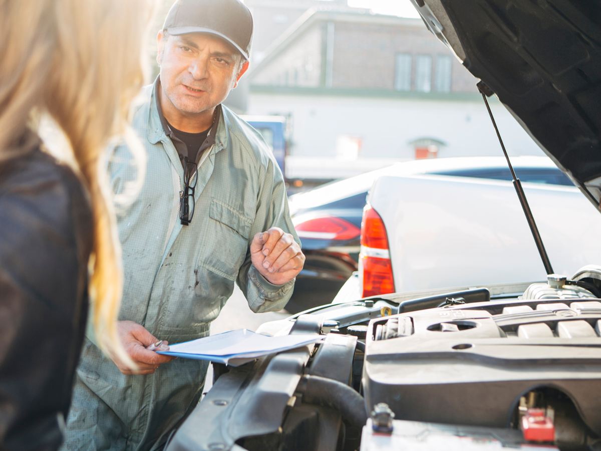 Mechaniker mit Klemmbrett erklärt einer Kundin am offenen Motorraum die Reparatur